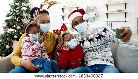Portrait of happy African American family in masks taking pictures on smartphone in cozy christmassy decorated home. Dad in santa hat taking selfie photo on cellphone with kids and wife