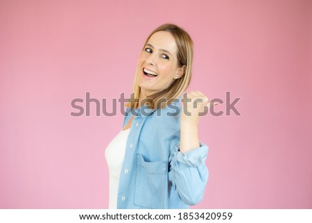 Beautiful young woman wearing denim shirt standing over isolated pink background pointing thumb up to the side smiling happy with open mouth