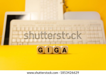 The word giga written with real wooden dices in front of a yellow background with computers and keyboard