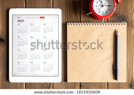 a tablet with an open calendar for 2021 year, a red alarm clock, and a craft paper notebook on a wooden boards table background Royalty-Free Stock Photo #1853450608