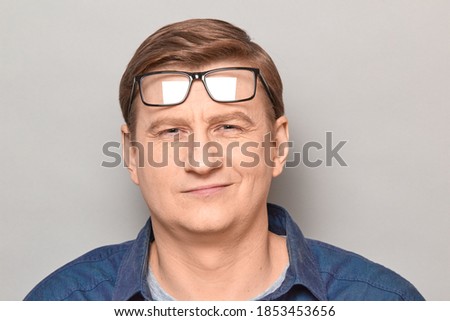Studio close-up portrait of happy blond mature man with glasses raised on forehead, smiling slightly, with positive expression on face, looking optimistic and confident. Headshot over gray background