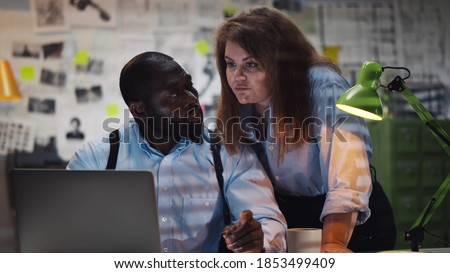 African man with woman colleague looking at laptop screen in police office. Diverse detectives coworkers analyzing data and discussing criminal case in agency office