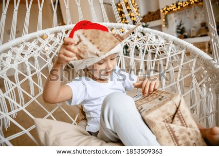 A four-year-old girl in a Santa hat sits in a wicker chair against the background of a Christmas tree and unwraps a gift
