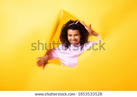 happy african american woman looking at camera through hole in ripped paper on yellow background