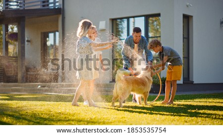 Smiling Father, Daughter, Son Play With Loyal Golden Retriever Dog, Spraying Each other with Garden Water Hose. On a Sunny Day Family Having Fun Time Together Outdoors in Backyard. Royalty-Free Stock Photo #1853535754