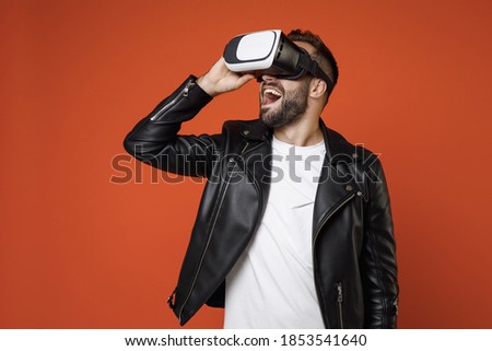 Excited cheerful young bearded man 20s wearing casual basic white t-shirt, black leather jacket standing watching in vr headset gadget isolated on bright orange colour background studio portrait