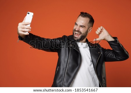 Funny young bearded man 20s wearing basic white t-shirt black leather jacket doing selfie shot on mobile phone pointing thumb on himself isolated on bright orange colour background studio portrait