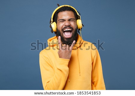 Funny young african american man guy in casual yellow streetwear hoodie isolated on blue wall background studio. People lifestyle concept. Listen music with headphones depicting heavy metal rock sign