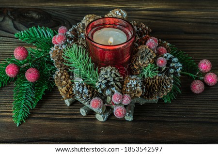 Candlestick with a lit candle in the center of a natural Christmas composition of branches of a Christmas tree, cones, artificial berries on a wooden background