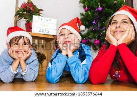 Mother and two kids lying on the floor with their hands on their chins wearing Christmas hats and with a Christmas tree and a light box in the background
