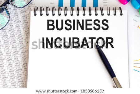 In the notebook is the text of BUSINESS INCUBATOR, next to the pen, glasses, reports and graphs.