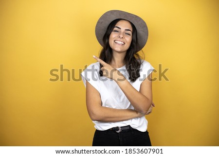 Beautiful woman wearing casual white t-shirt and a hat standing over yellow background smiling and pointing with hand and finger to the side