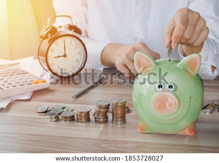 Female's hand put coin on green piggy bank and stack coins on wood table. for saving money with business stuff, Business and finance concept