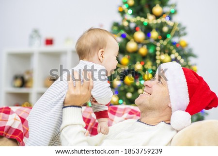 Young dad in a santa hat holds his son in his arms against the background of a Christmas tree at home