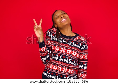 Young beautiful African American woman wearing Christmas sweater, against red background smiling with happy face winking at the camera doing victory sign. Number two.