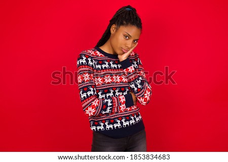 Very bored Young beautiful African American woman wearing Christmas sweater, against red background holding hand on cheek while support it with another crossed hand, looking tired and sick,