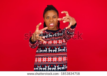 Young beautiful African American woman wearing Christmas sweater, against red background Shouting frustrated with rage, hands trying to strangle, yelling mad.