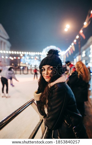 Evening portrait of a cute girl in warm clothes standing near the rink on background of the evening Christmas fair looks at camera and smiles. Christmas outdoors photos of a beautiful lady