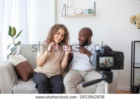 A cheerful multinational couple is streaming live video to their blog. Curly future mommy shows the audience the baby socks. Family, pregnancy and technology concept.