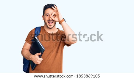Young hispanic man wearing student backpack holding binder smiling happy doing ok sign with hand on eye looking through fingers 
