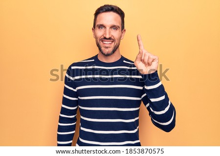 Young handsome man wearing casual striped sweater standing over isolated yellow background smiling with an idea or question pointing finger up with happy face, number one