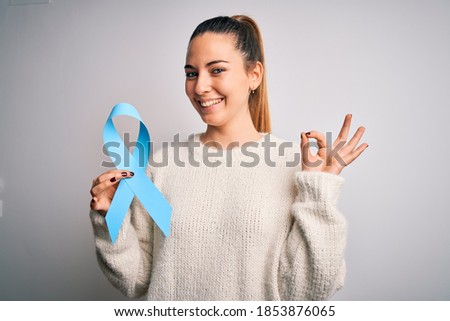 Young beautiful blonde woman holding blue cancer ribbon over isolated white background doing ok sign with fingers, excellent symbol