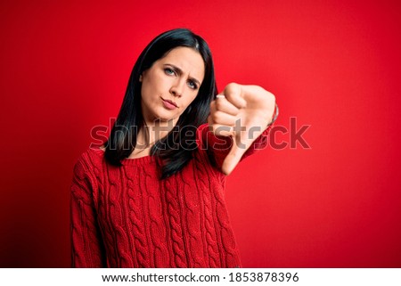 Young brunette woman with blue eyes wearing casual sweater over isolated red background looking unhappy and angry showing rejection and negative with thumbs down gesture. Bad expression.