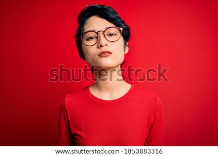 Young beautiful asian girl wearing casual t-shirt and glasses over isolated red background Relaxed with serious expression on face. Simple and natural looking at the camera.
