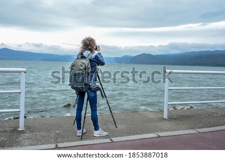 Young female photographer with backpack standing on riverbank and taking a photo of river and mountain with digital camera, back view