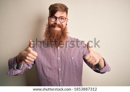 Handsome Irish redhead business man with beard wearing glasses over isolated background success sign doing positive gesture with hand, thumbs up smiling and happy. Cheerful expression and winner 
