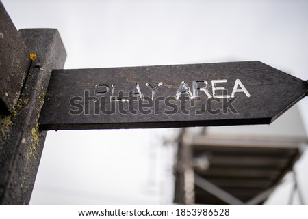 Up close view of a wooden pole with indications pointing out the way to the play area, with a cloudy sky and a blurry structure as background