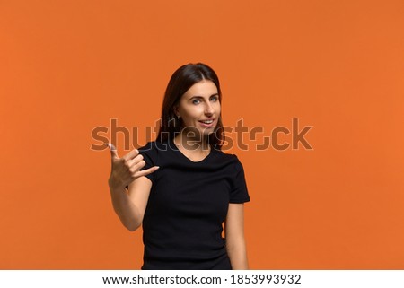 Enjoy distant communication. Positive talkative caucasian woman in black t-shirt makes telephone gesture, smiles pleasantly. Isolated over an orange background.