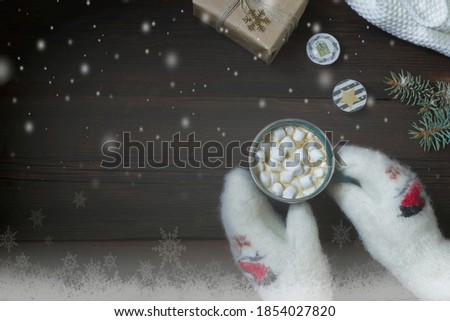 Female hands in white woolen mittens hold a cup of hot drink with marshmallows. New Year's gift, candles on a dark wooden background with imitation of snowflakes. Christmas New Year card. Copy space.