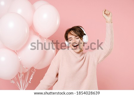 Funny young woman in knitted casual sweater isolated on pastel pink background. Birthday holiday party people emotions concept. Celebrating hold air balloons listening music with headphones dancing
