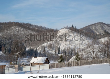 Winter, frosty day in a village located among snow-capped mountains. The mountain slopes are covered with coniferous forest.