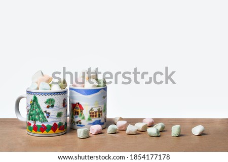 two christmas mugs with chocolate and colored bonbons, on table decorated with more marshmallows and white background