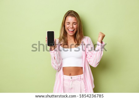 Image of excited blond girl winning something, showing app achievement on mobile phone screen, saying yes and triumphing, standing over green background