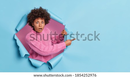 Shocked dark skinned woman with curly hair points away on blank space sees something incredible and breathtaking wears casual knitted sweater breaks through paper background. Omg look there. Royalty-Free Stock Photo #1854252976