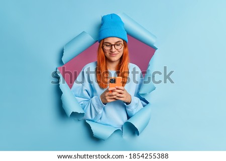 Redhead pleased young Caucasian woman uses mobile phone types sms message surfs in social networks wears blue hat and sweatshirt poses in ripped hole through paper background. Chatting online