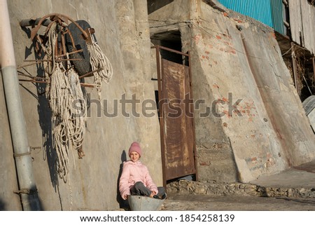 Nice girl in a pink jacket sits in a basin near a concrete wall in a fishing village, hang anchors on the 