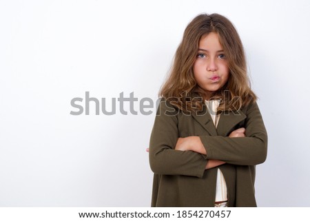 Picture of angry Young beautiful Caucasian little girl standing against white background crossing arms. Looking at camera with disappointed expression.