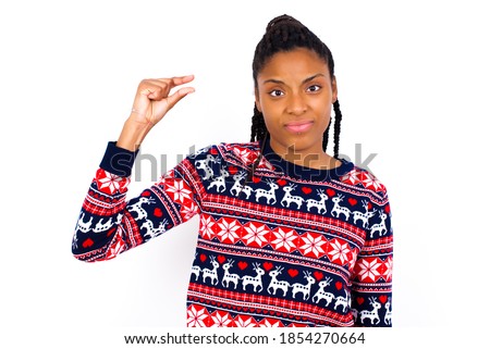 Upset Excited Young beautiful African American woman wearing Christmas sweater, against white wall shapes little gesture with hand demonstrates something very tiny small size. Not very much