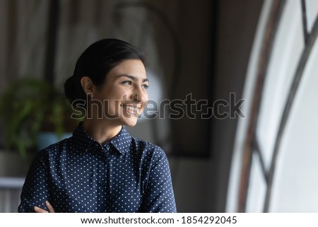 Close up smiling dreamy Indian businesswoman looking to aside through window, standing in office with arms crossed, happy female employee intern thinking about future, visualizing, business vision