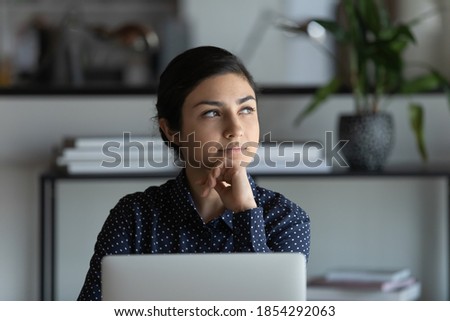 Close up thoughtful Indian businesswoman touching chin, using laptop, sitting at desk in modern office, pensive employee pondering difficult task, developing project or business strategy