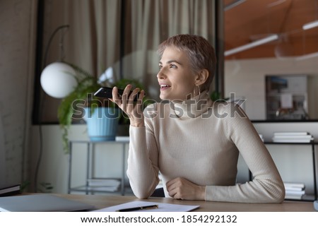 Smiling businesswoman recording voice message on phone, sitting at desk in office, satisfied female employee holding smartphone near face, chatting online, making call, talking by speakerphone
