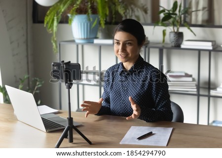 Successful Indian businesswoman recording webinar, using smartphone on tripod, sitting at desk in office, smiling young employee business coach teacher influencer shooting video for blog, speaking Royalty-Free Stock Photo #1854294790