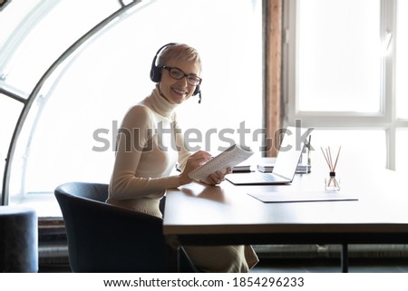 Portrait smiling businesswoman wearing headset sitting at work desk, looking at camera, happy confident female employee manager in headphones writing notes, watching webinar, consulting client online