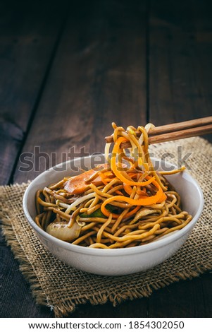 Chopsticks take chinese vegetarian noodles, served in a white bowl on rustic wooden background, vertical stock photo