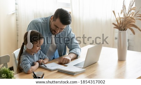 Caring father helping little daughter with school homework, sitting at table at home, child schoolgirl wearing headphones studying online, using laptop, dad checking tasks, homeschooling concept Royalty-Free Stock Photo #1854317002