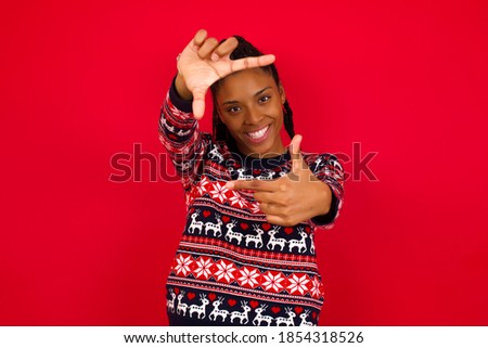 Young beautiful African American woman wearing Christmas sweater against white wall, making finger frame with hands. Creativity and photography concept.